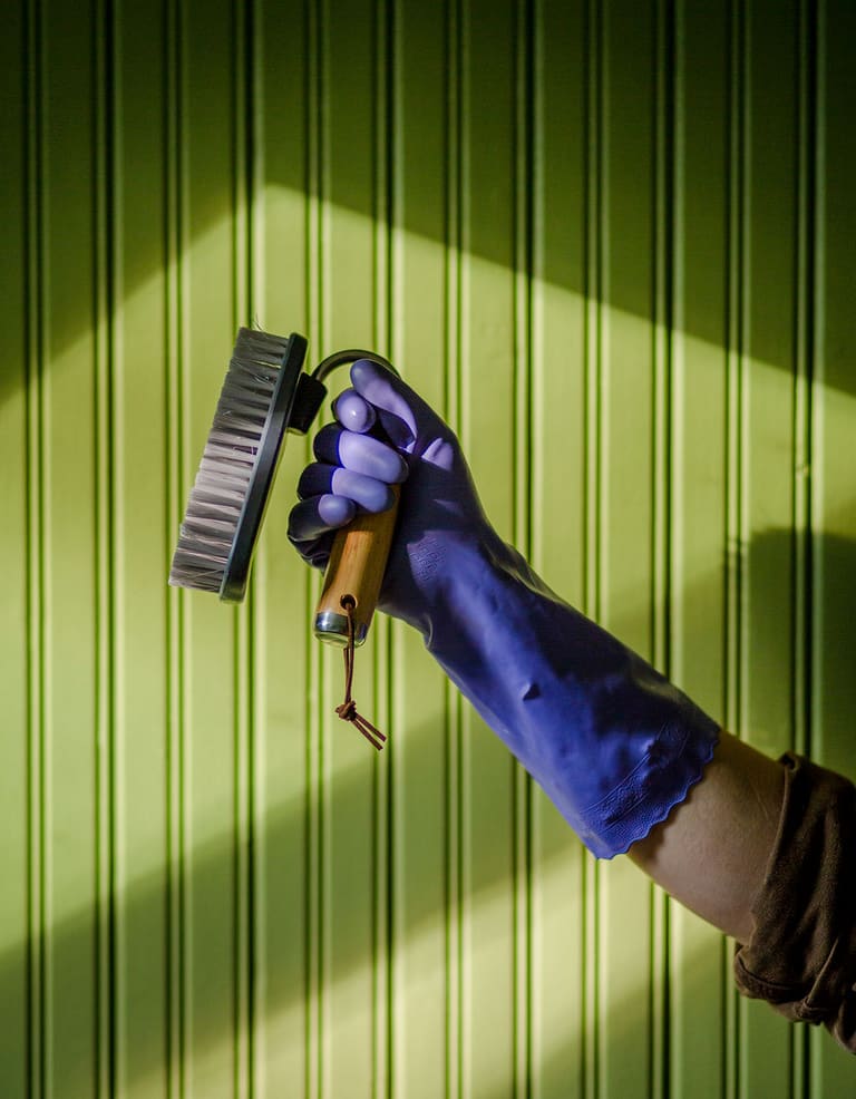 A purple-gloved hand holding a wooden scrub brush against green beadboard, lit by raking sunlight