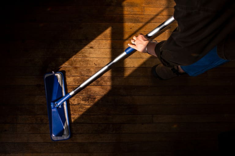 Looking down at a flat mop being pushed across wide pine floorboards through a stripe of sunlight