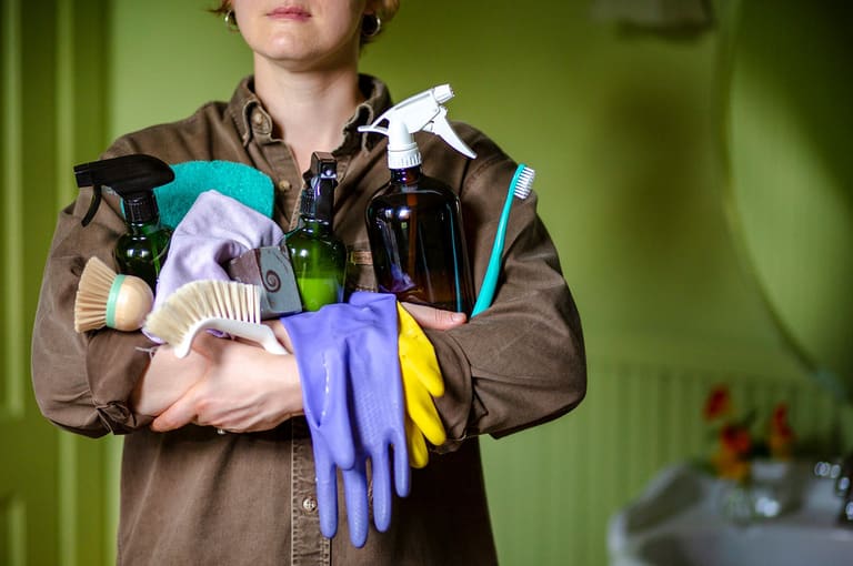An armful of supplies — amber spray bottles, folded cloths, scrub brushes and rubber gloves — carried against a green wall