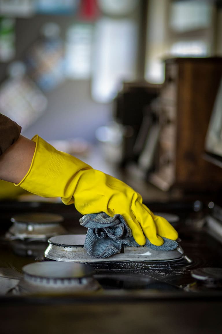 A yellow-gloved hand wiping soapy water off a gas burner ring on a stovetop
