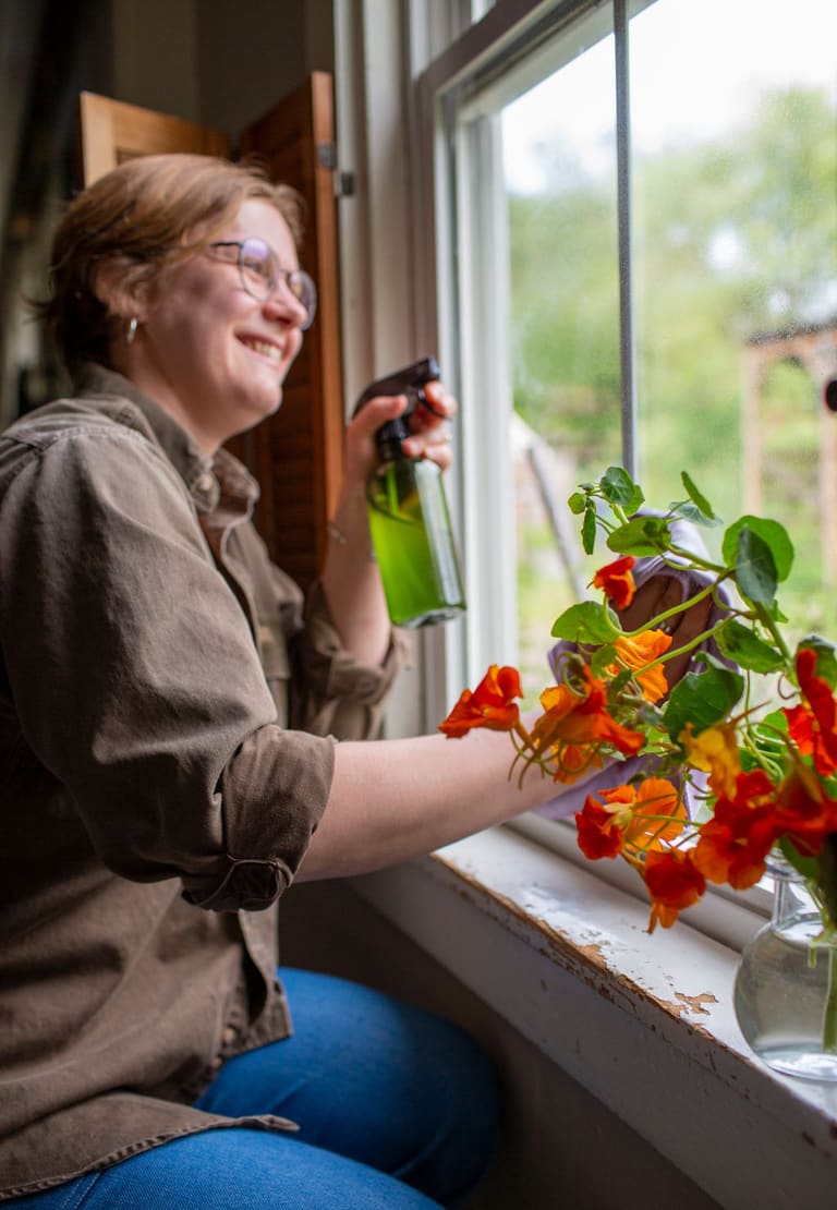 Wiping down an old window sash with a spray bottle and cloth, orange nasturtiums flowering on the sill